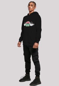 Fekete kapucnis póló "CENTRAL PERK" logóval piros és zöld színben, két fehér kávéscsészével. Elülső kenguruzsebbe és bordás mandzsettákba kerül.