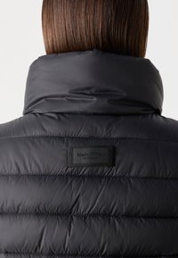 Doudoune noire avec un col montant, présentant une texture matelassée et une étiquette de marque rectangulaire au dos. Matériau lisse et brillant.