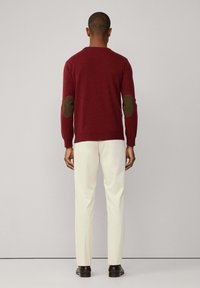 Muškarac u bordo puloveru s tamnim laktovima, kremastim hlačama i tamnim cipelama, stoji okrenut prema jednostavnom svijetlosivom zidu.