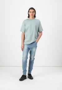 Svijetloplavi pamučni T-shirt, opuštenog kroja, kratkih rukava. Uparen sa svijetloplavim trapericama i crnim tenisicama. Jednostavna pozadina.