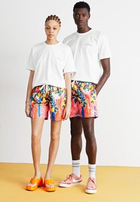 Witte t-shirts, kleurrijke zwembroeken met patronen in bloemen- en streepdessin. De shorts zijn vastgemaakt met een koord. Heldere oranje schoenen.