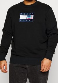 Černá mikina s žebrovanými manžetami a lemem, s obdélníkovým vyšívaným logem v bílé, modré a kárované barvě.