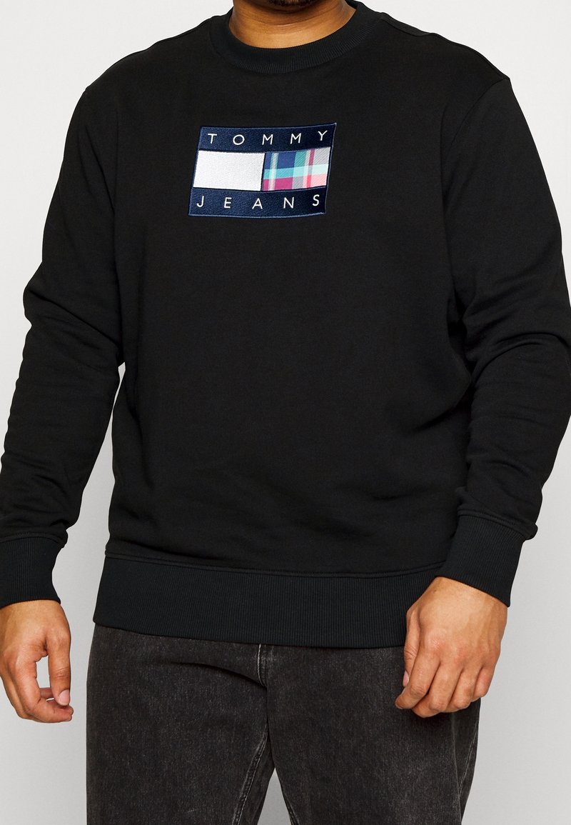 Černá mikina s žebrovanými manžetami a lemem, s obdélníkovým vyšívaným logem v bílé, modré a kárované barvě.