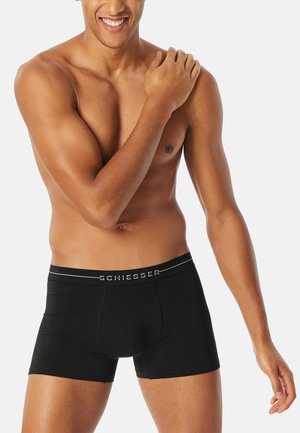 Lächelnder, oberkörperfreier Mann mit mittlerem Hautton, der schwarze Schiesser Boxershorts trägt und mit seiner rechten Hand seine linke Schulter hält.