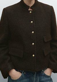Brown wool jacket with a high collar, gold buttons, and two front pockets. The fabric has a soft texture and a boxy, cropped design.