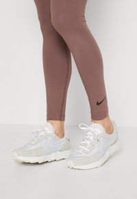 Nike tenisice s bijelo-svijetlosivom shemom boja, kombinacijom materijala uključujući mrežicu i antilop, s teksturiranim gumastim potplatom i vezicama.
