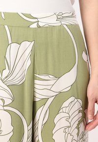 Tissu vert avec un grand imprimé floral blanc porté par une personne en haut blanc côtelé, montrant une partie du bras et de la main reposant sur le côté.