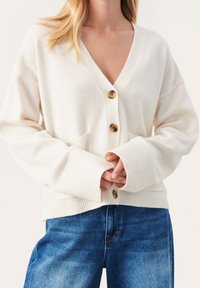 Witte gebreide cardigan met een diepe V-hals, vier bruine knopen, geribde manchetten en opgestikte zakken aan de voorkant, gecombineerd met blauwe denim jeans.