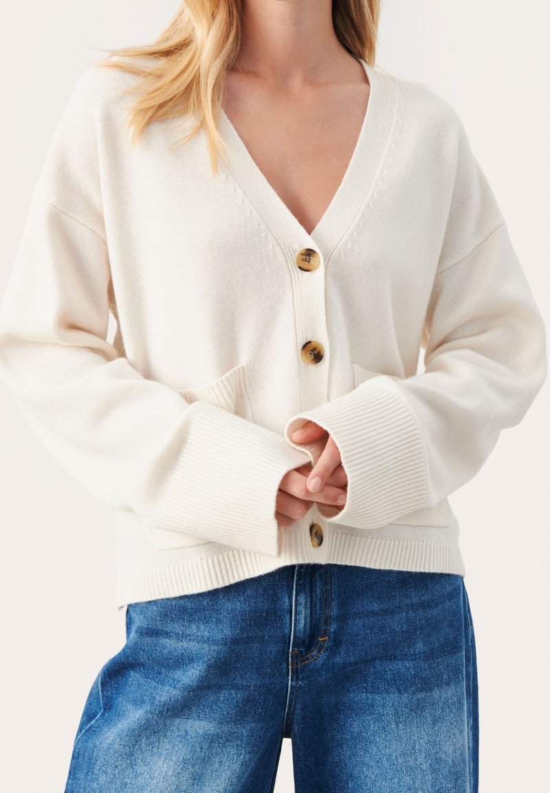 Witte gebreide cardigan met een diepe V-hals, vier bruine knopen, geribde manchetten en opgestikte zakken aan de voorkant, gecombineerd met blauwe denim jeans.