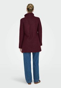 Burgundijas vilnas mētelis ar augstu apkakli un garām piedurknēm, kombinēts ar platām zils džinsu biksēm. Minimalistiskais dizains, teksturēta apdare.