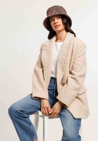 Cappotto beige oversize con texture e maniche ampie, abbinato a jeans blu a vita alta. Il modello indossa un cappello a secchiello con un motivo multicolore.