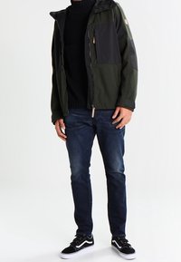 Zeleno-černá bunda na zip s kapucí, vybavená přední náprsní kapsou, spolu s tmavými džíny a černými botami s bílou podrážkou.