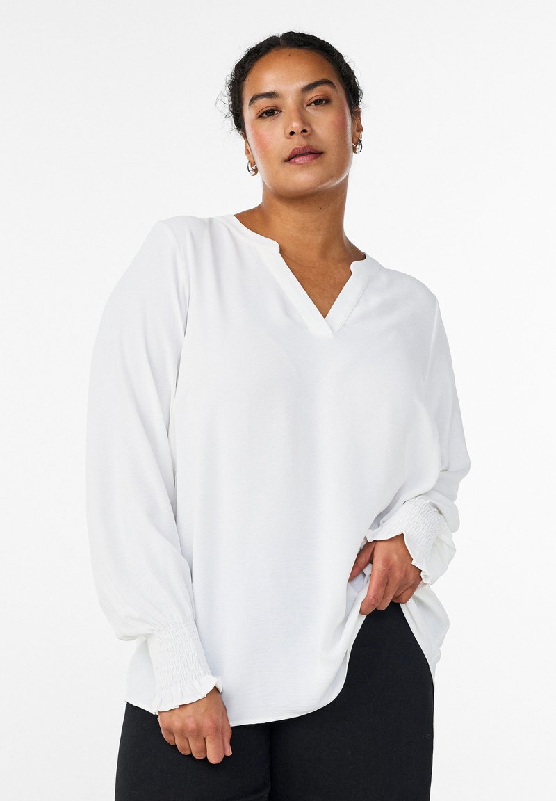 Mujer con cabello rizado y oscuro, vistiendo una blusa blanca de manga larga con puños fruncidos y pantalones negros, de pie frente a un fondo liso.