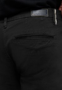 Zwarte broek met een leren label met "GEEN EXCUSES," een achterzak met subtiele stiksels en een zachte stoftextuur.