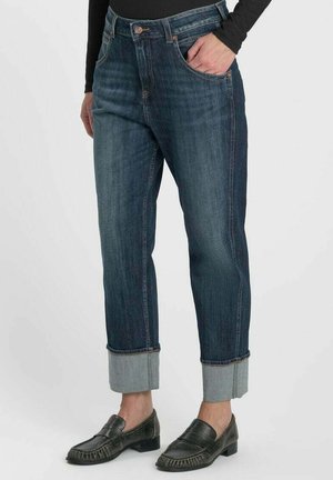 Personne portant un jean bleu foncé à revers et des mocassins en cuir noirs, avec une main dans la poche, debout sur un fond clair.