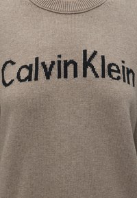 Pull beige en maille avec texte logo noir "Calvin Klein" sur la poitrine et col rond côtelé.