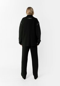 Chaqueta negra de gran tamaño con hombros caídos y detalles metálicos, combinada con pantalones de pierna ancha a juego. Tejido suave con un corte relajado.