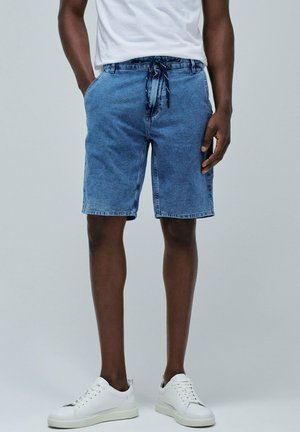 Shorts de mezclilla en azul claro, con una cinturilla con cordón, bolsillos laterales y un corte holgado, combinados con zapatillas blancas.