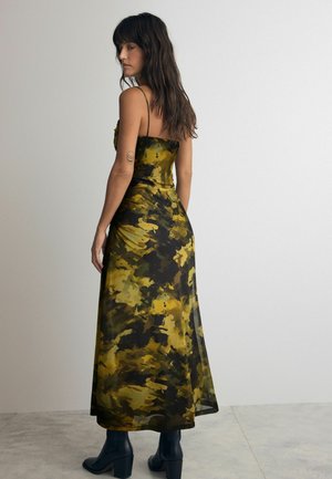 Vestido maxi con estampado de camuflaje en verde y amarillo, con tirantes finos y un corsé ajustado, que fluye hacia un dobladillo ligeramente transparente. Llevado con botas negras.