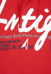 Tissu rouge avec texte imprimé en blanc « Antigua » et détails gris incluant « Heritage Quay Ports Docking ». Comprend une étiquette avec la taille et les instructions d'entretien.