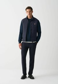 Navy blue puffer vest with vertical quilted pattern, zippered front, and round logo patch, worn over a matching navy sweater and trousers.