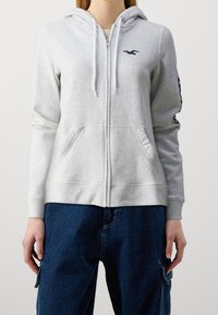 Sudadera con capucha gris claro con cremallera, hecha de tela suave, con bolsillo frontal, capucha con cordón y logo azul marino en el pecho izquierdo y la manga.