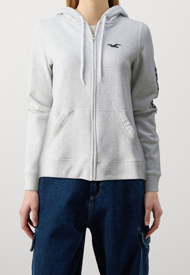 Sudadera con capucha gris claro con cremallera, hecha de tela suave, con bolsillo frontal, capucha con cordón y logo azul marino en el pecho izquierdo y la manga.