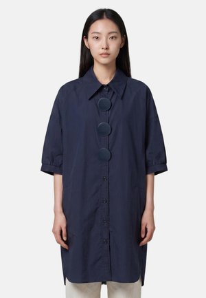 Giovane donna con lunghi capelli neri che indossa un vestito camicia oversize blu navy scuro con grandi bottoni rotondi e maniche alla lunghezza del gomito.
