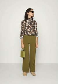 Blusa com padrão abstrato em castanho e creme, calças largas verdes-oliva, óculos de sol circulares e uma pequena malinha quadrada verde.