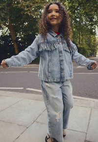 Hellblaue Jeansjacke mit Fransen, kombiniert mit ausgefransten, weit geschnittenen Jeans. Korksandalen runden das Outfit ab.