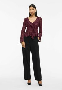Blouse en dentelle bordeaux avec des détails volantés et des manches longues, associée à un pantalon tailleur noir taille haute et des talons noirs.