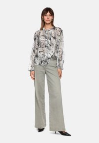 Durchsichtige Bluse mit monochromem Stadtbilddruck, langen Ärmeln und einem rüschierten Ausschnitt, kombiniert mit grauen, weit geschnittenen Hosen und schwarzen, spitzen Schuhen.