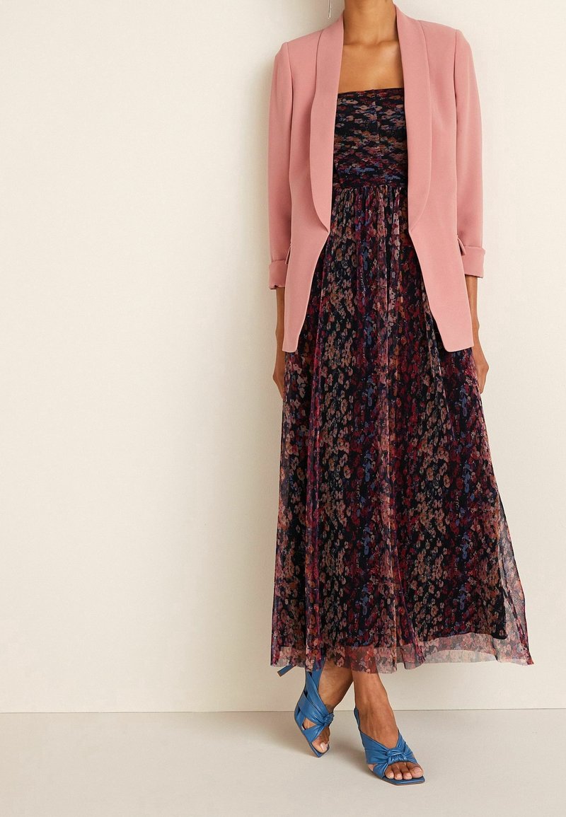 Largo vestido floral en colores oscuros, con tejidos en capas y un ligero brillo, combinado con una chaqueta rosa. Zapatos azul de punta abierta añaden contraste.