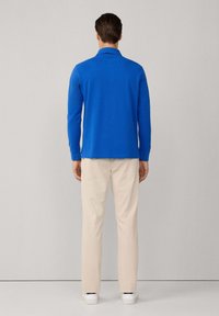 Mann, der mit dem Rücken zur Kamera steht, trägt ein langärmliges blaues Hemd, beigere Hosen und weiße Sneakers vor einem einfarbigen Hintergrund.