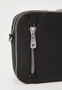 Bolso de cuero negro con un acabado texturizado, que presenta cremalleras y herrajes en plata. Forma rectangular compacta con un bolsillo lateral con cremallera.