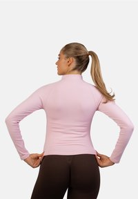 Langærmet bluse i lys rosa med høj krave og glat tekstur, parret med brune leggings, vist fra ryggen.