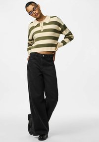 Osoba ubrana w ciemne szerokie spodnie, czarne buty oraz zielono-kremowy krótki sweter w paski z kołnierzykiem i guzikami, pozująca z rękami na biodrach.