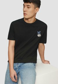 Czarny bawełniany t-shirt z krótkimi rękawami, okrągłym dekoltem oraz haftowanym wzorem w niebiesko-białe na lewej piersi. Noszony z jasnoniebieskimi dżinsami.