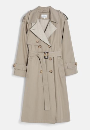 Femme portant un trench beige, un pull à col roulé crème, un pantalon beige à jambes larges et des chaussures marron pointues, debout les mains dans les poches.