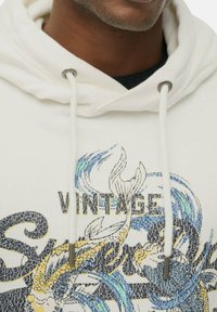 Sudadera blanca con un estampado gráfico que presenta elementos en azul y amarillo, cordones con puntas de metal y una textura suave.