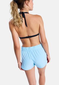 Woman with blonde hair in a ponytail wearing a black halter bikini top and light blue swim shorts, facing away against a white background.