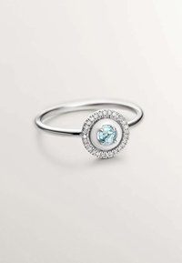 Anillo de plata con un diseño circular que presenta una piedra preciosa azul central rodeada por un halo de pequeños cristales claros. Textura de banda suave.