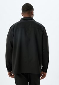 Camisa negra de manga larga con un ajuste relajado, que presenta un puño abotonado, cuello con solapa y una textura suave y mate.
