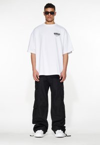 T-shirt bianca oversize con un logo nero vicino al petto sinistro, abbinata a pantaloni cargo neri e sneaker bianche.