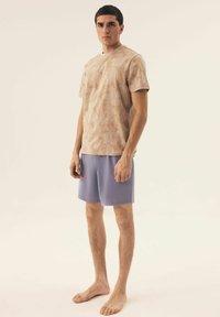 Jasnopomarańczowy t-shirt w wzory z krótkimi rękawami, zestawiony z jednolitymi, lawendowymi szortami. Tkanina wydaje się miękka i wygodna. Model stoi na boso.