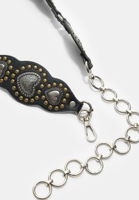 Črni usnjeni ovratnik z metalnimi zatiči in srčkastimi poudarki, z srebrno verigo in sponko. Teksturirana površina z drznih detajli.