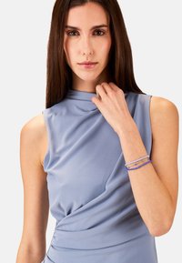 Ljusblå figurnära klänning med hög halsringning och mjukt drapering. Armbandet har flera trådar av blå pärlor. Slät tygtextur.