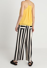 Top sin mangas amarillo con borde de encaje, combinado con pantalones de pierna ancha a rayas en blanco y negro. El conjunto presenta un corte relajado y una tela ligera.