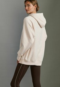Un hoodie rose clair avec une capuche à cordon de serrage, coupe décontractée et poignets côtelés. Associé à des leggings marron avec des rayures latérales blanches et roses.