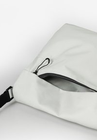 Lichtgrijze tas met een gladde buitenkant, voorzien van een diagonale ritsluiting, een verstelbare zwarte schouderriem en een trekkoordaccent.